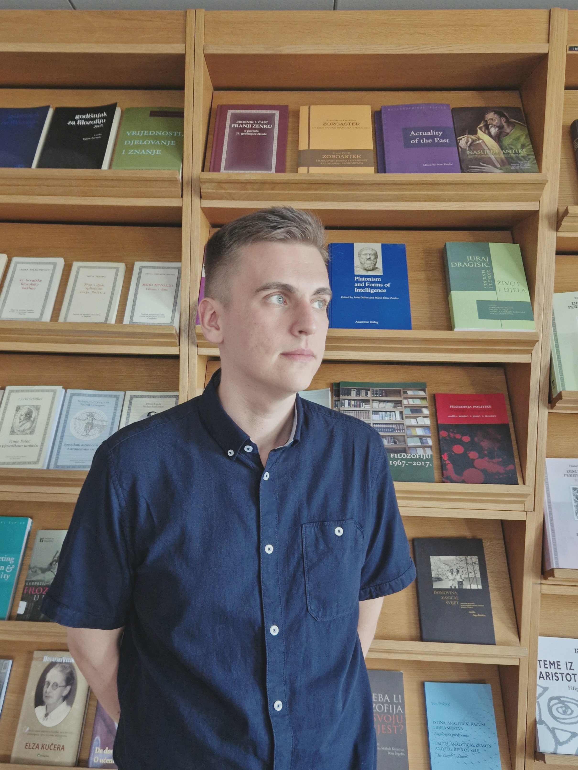 Photo by Mia Biturajac. Me, male, 30s, white, blue eyes, short light brown hair swept to the side, dark blue button-up shirt, average build, in front of a bookshelf with philosophy books.